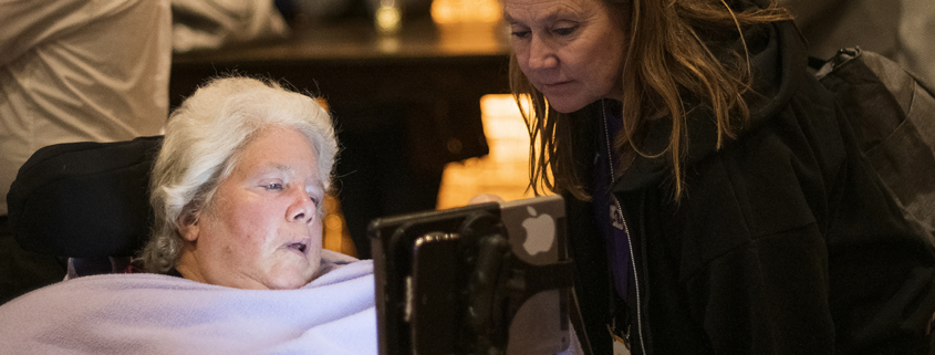 An older woman with short gray hair and covered in a blanket looks at her assistive technology screen, with another woman standing beside her and also viewing the screen