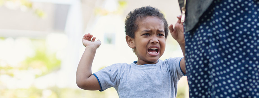 A 3 or 4 year old boy is upset at his mother, standing nearby, and raising his arms as he throws a temper tantrum. They are outside.