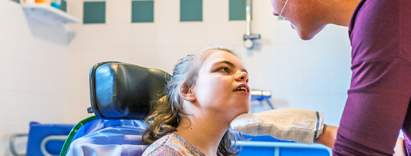 A caregiver lifts the chin of a young girl with disabilities who is sitting in a patient chair.