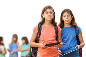 Two sisters with glasses, backpacks, and tablets stand against a white background, with other students unfocused behind them.