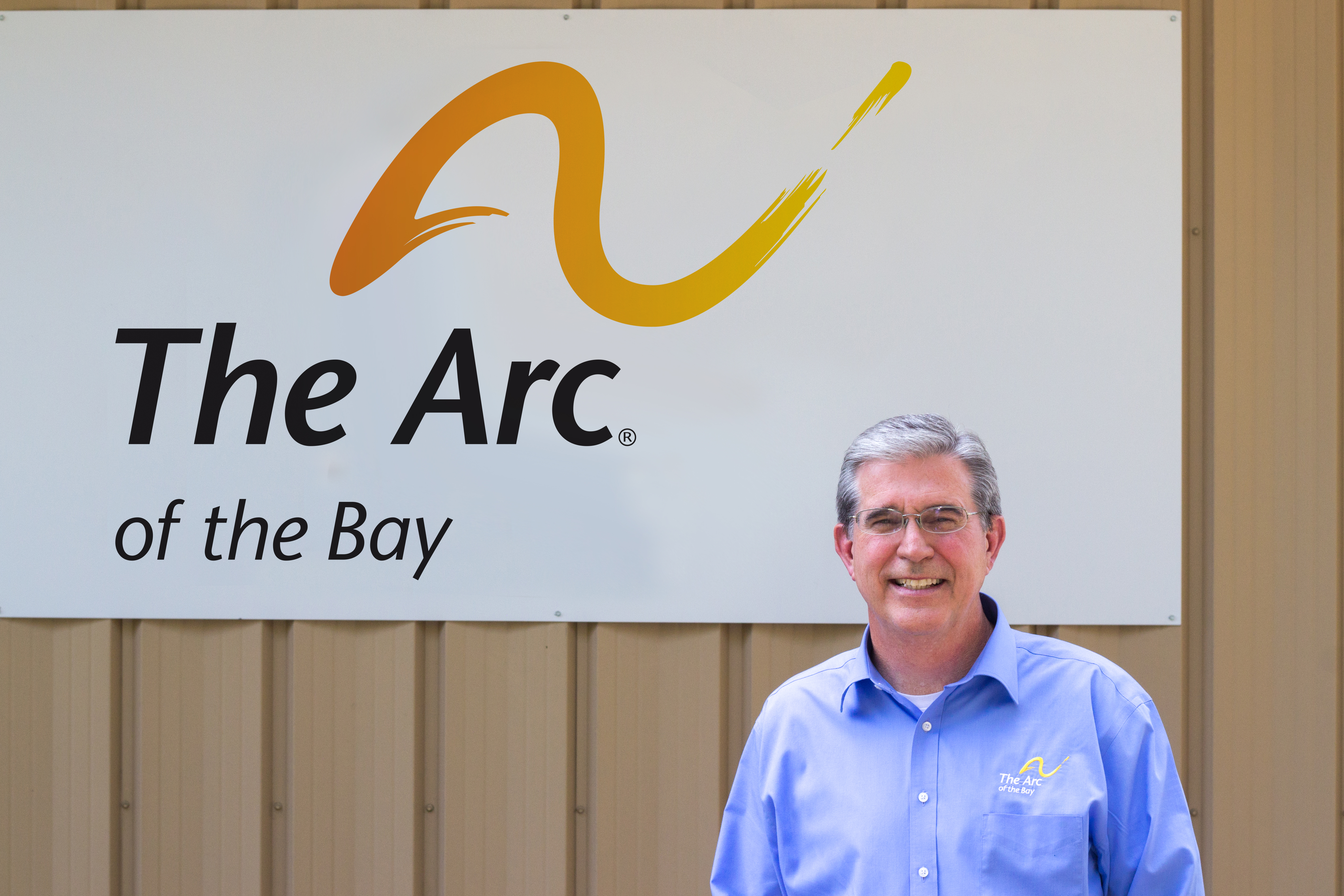 Ron Sharpe, Executive Director of The Arc of the Bay, stands smiling in front of a sign with the chapter's name on it. 