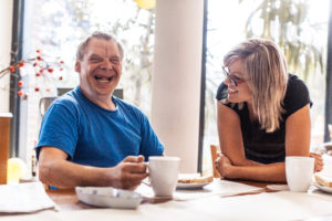 An adult man with gray hair sits smiling and laughing as he holds a cup of coffee, next to his support professional - a woman with glasses who looks at him smiling. 