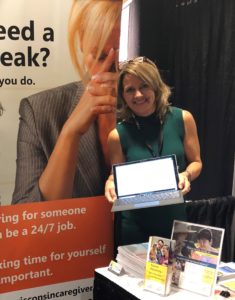 The Arc Wisconsin State Director Lisa Pugh stands in front of an exhibiting booth smiling and holding a computer. 