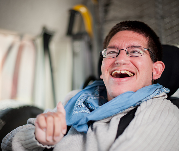 Man with a disability smiling and seated. He has glasses and is wearing a blue bandana around his neck