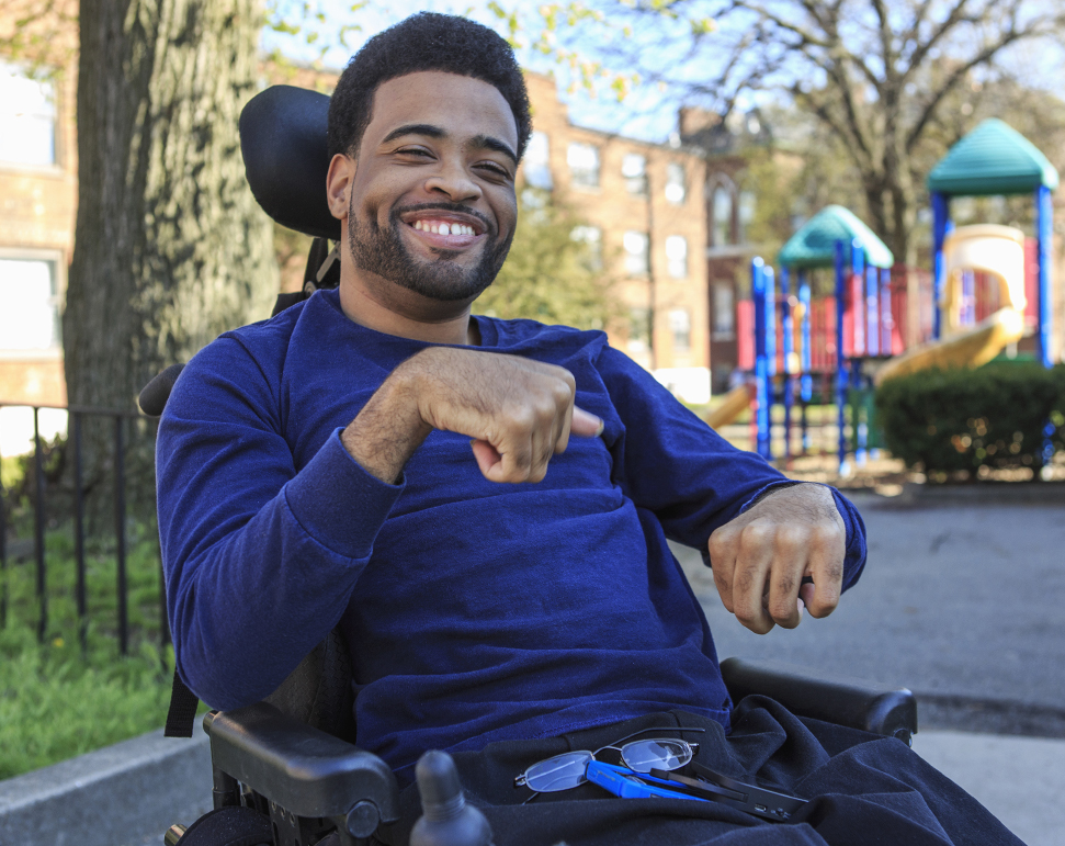 Smiling man with a disability in a wheelchair at a park, with a playground in the background