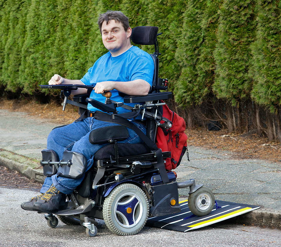 Smiling man with disability in motorized wheelchair riding down a sidewalk ramp