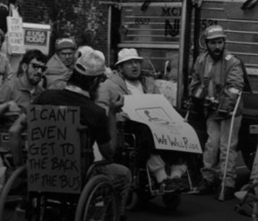 A historical black and white photo of activists with mobility aids. The back of a man's wheelchair reads "I can't even get to the back of the bus"