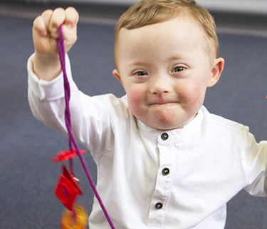 Toddler with Down syndrome sits on a floor and proudly holds up his toy necklace