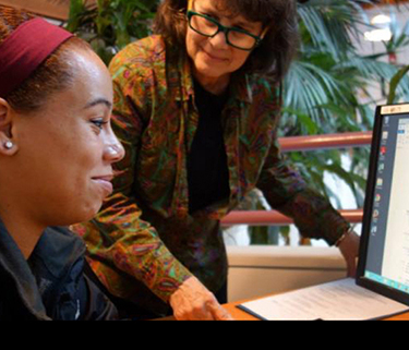 A woman smiles as she looks at a computer, while another woman stands next to her smiling