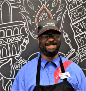 Kevin stands smiling in front of a black chalk wall with drawings on it, wearing his employee name tag, an apron, and a hat. 