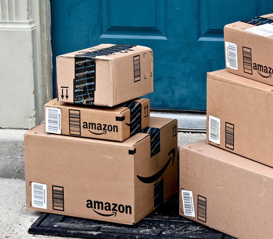 Amazon boxes of various sizes sit, stacked, in front of a doorstep.