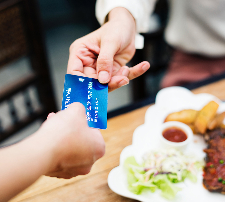 A credit card is passed between two people, with food out of focus on a table in the background