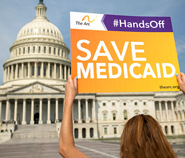A woman stands facing the Capitol Building in DC, with her back to the camera, holding a sign that says "#HandsOff: Save Medicaid".