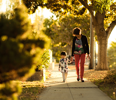A woman and young child walk down a sun-filled sidewalk holding hands