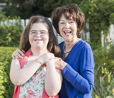 A teenage girl with Down syndrome standing in a yard in front of a white fence with an older family member. The woman is laughing, with her hands on her granddaughter's shoulders. They are both looking at the camera.