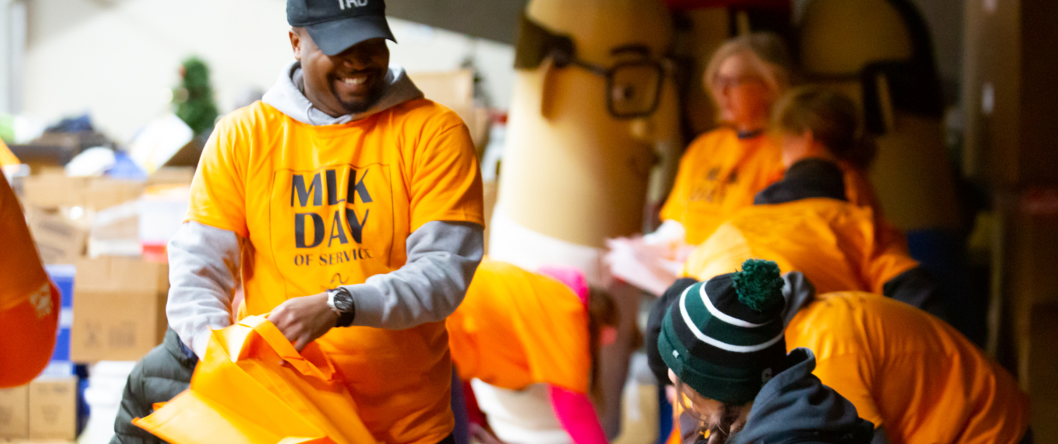 A man wearing an orange "MLK Day of service" shirt and black hat smiles at another event participate, as people also wearing orange shirts stuff boxes for donation behind them