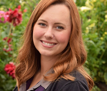 Headshot of smiling woman with red hair in front of flowers and bushes