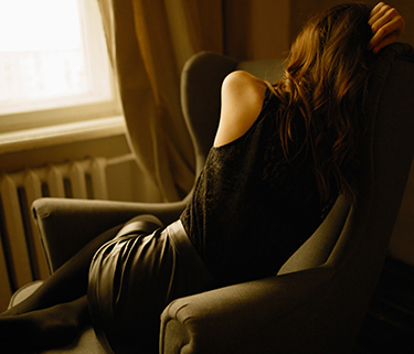A woman faces away towards a window, sitting in a chair with sad body language.