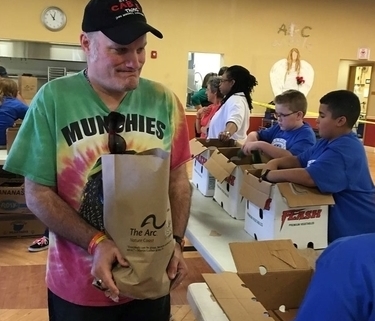 A volunteer in a tie dye shirt holds a paper bag of food donations