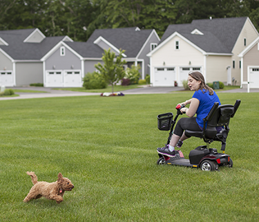A woman in a motorized chair plays with a small dog on a grassy field in front of a community of houses