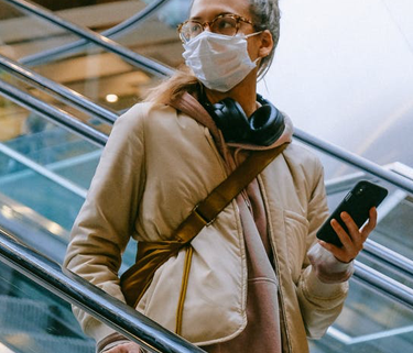 Woman on escalator wearing a face mask; she's holding a cell phone in one hand and her suitcase handle in the other