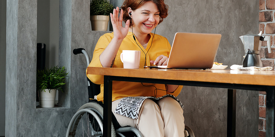 A woman sits at a table on a virtual call, smiling and waving at her computer.