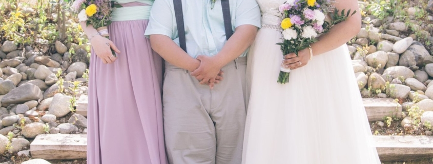 Three people standing outside at a wedding. On the left is a bridesmaid wearing a strapless purple dress and holding a bouquet of yellow and purple flowers. In the middle is a man with Down syndrome. he is wearing grey pants with blue suspenders, a light blue short sleeve button up shirt and is wearing a purple flower pinned to his shirt. On the right is the bride, dressed in a white, lacey, bridal gown and holding a bouque of purple and yellow flowers. They are standing on gravel and behind them are stairs leading up to a stone and log cabin.