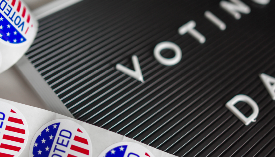 A close up image of red,white and blue stickers that have stars and stripes on them with the words "I voted" in the middle. Next to the stickers is a black board with white letters that say "vote"