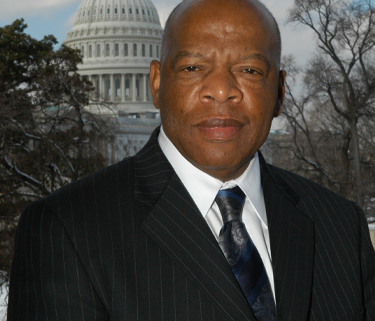 A man wearing a black pinstripe suit jacket, white dress shirt and blue floral silk tie posing. In the background is a photo of trees and the Capitol building.