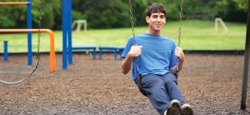 A young man swinging at a playground. He has short brown hair and is wearing a blue t-shirt and blue sweat pants. In the background is playground equipment and past the playground is a green field with soccer goals. There is a forest after the soccer field.
