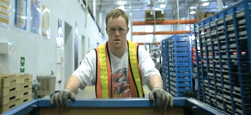 A man is in a factory. He is wearing a white t-shirt with a photo of racing cars on it. He is wearing an orange and yellow reflective safety vest and goggles. He has gloves on and is pushing a large cart.
