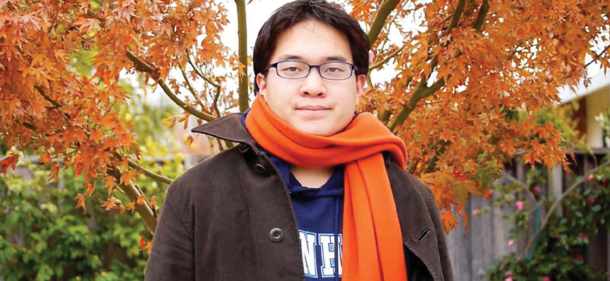 A photo of a person from the chest up. He is wearing a black jacket over a blue t-shirt and has a bright orange scarf wrapped around his neck. He has short hair and glasses. He is standing outside under a tree with bright orange leaves.