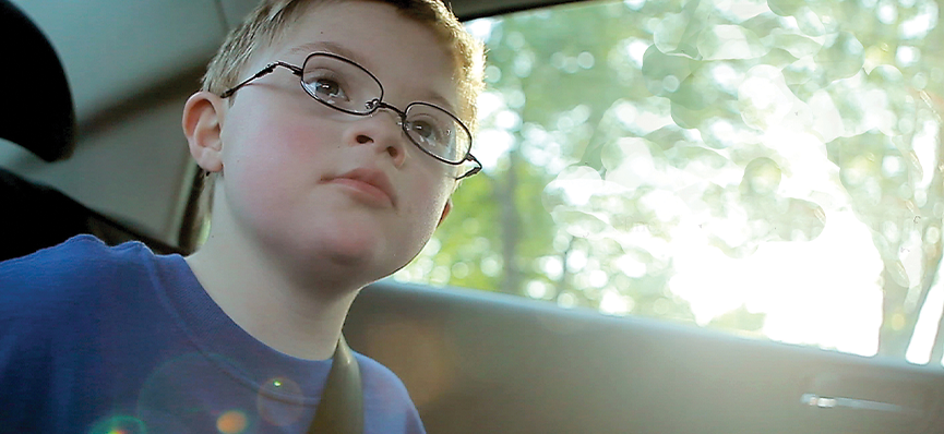 A young boy sits in the back of the car. He has short blonde hair and glasses. He is wearing a blue t-shirt. Next to him is the the car window. In the background of the window are blurred trees and bright sun.