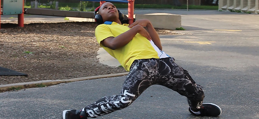 A man at a park leans way back in a dance move. He is wearing head phones and has on a bright yellow shirt and black pants with a white print on them.