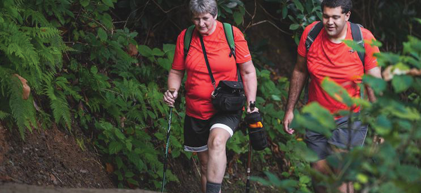A older woman and a young man are hiking on a hiking trail. There are high embankments with ferns and dirt on the left side. The woman has short grey hair. She is wearing a red short sleevel t-shirt and black shorts and holding walking sticks. She is wearing a backpack and has another satched across her front. The young man has short brown hair. He is also wearing a short-sleeve red t-shirt and grey shorts and is wearing a backpack.