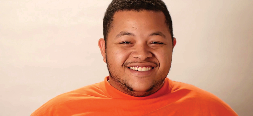 A photo of a man from the shoulders up. He has short brown hair and brown facial hair. He is wearing an orange shirt and smiling.