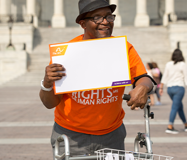 A man uses a walker with a basekt it on it holding the walker with one hand and a sign in the other hand. He is standing in front of the Capitol building. The sign has The Arc logo in the top left corner and is blank in the middle.