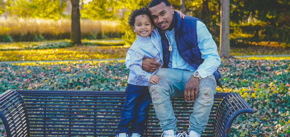A man sits at on the back of a park bench, his son stands on the park bench next to him and he has his arm around his sun. In the background, there are trees and leaves on the ground. The man is wearing light colored jeans with destruction on them and a long sleeve button up shirt. He has on a blue puffer vest and a gold chain and cgold watch. The young boy is wearing a long sleeve button up shirt and dark blue jeans.