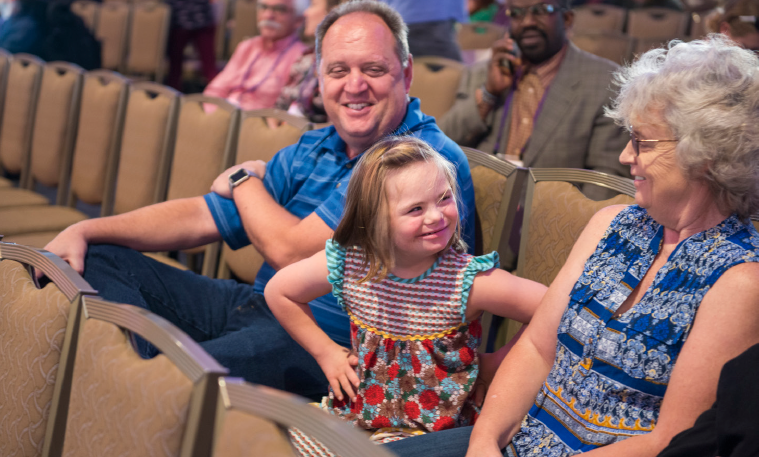 A young girl with Down syndrome sits in between a man and a woman in a conference room. She has her hands on her hips, looking athe woman and smiling. There are people sitting in random chairs in the background.