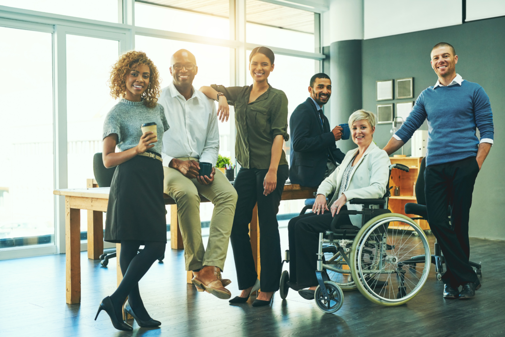 A diverse group of people dressed professionally are in an office. They are posing for the camera; ne person is in a wheelchair, two people sit on a table and the rest are standing.