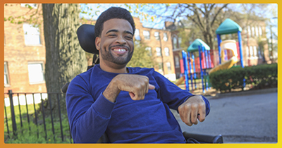 A photo of a man in a wheelchair in a park. He is wearing a blue long sleeve shirt and smiling.