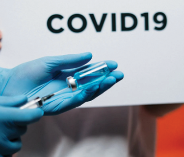 A gloved hand holding a vaccine vial, with the words COVID-19 in black on a board behind it.