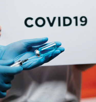 A gloved hand holding a vaccine vial, with the words COVID-19 in black on a board behind it.