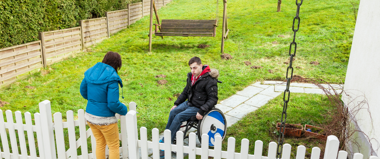 A man in a wheelchair exits a yard through a white picket fence. Behind him is a bench, and holding the gate open is a person in a blue jacket with yellow pants.