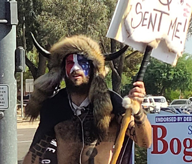 A photo of a man from the waist up. He is not wearing a shirt and has paint or tattoos on his body. He has face paint in red white and blue and is wearing a fur hat with tails coming down on either side. The hat has horns sticking out of it. He is holding a sign that says "Q Sent me"