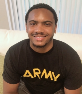 A young man sits smiling on a white couch with white blinds in the background. He is wearing a black shirt with the yellow word "ARMY" on it.