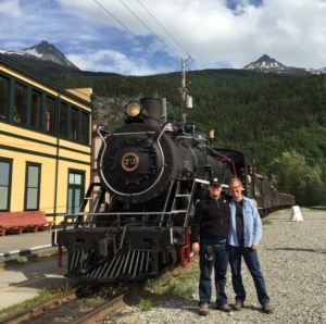 A father and adult son stand together, smiling, in front of a black train with green hills and mountains in the background. To the left of the train is a yellow building. 