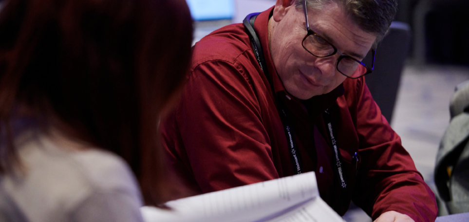 A man wearing a red shirt, glasses, and a blue lanyard that says "walmart" on it is looking at papers with another person.
