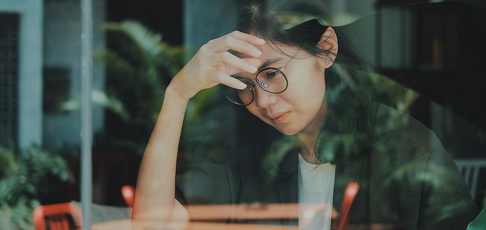 A photo taken of a woman through a window. She is sitting at a table with her hand on her forehead. She has long black hair and is wearing glasses and a jacket over a white shirt.
