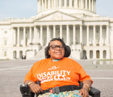 A woman sits in a motorized wheelchair with the US Capitol building in the background. She is smiling and wearing glasses, colorful floral pants, and an orange shirt with The Arc's logo that reads "Disability Rights are Human Rights"